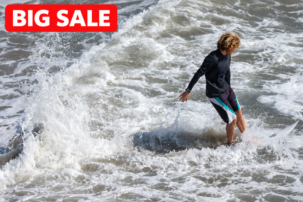 A surfer stands on a surfboard in foamy ocean water, wearing a black long-sleeve wetsuit top and black board shorts with a bright blue-green side stripe.
